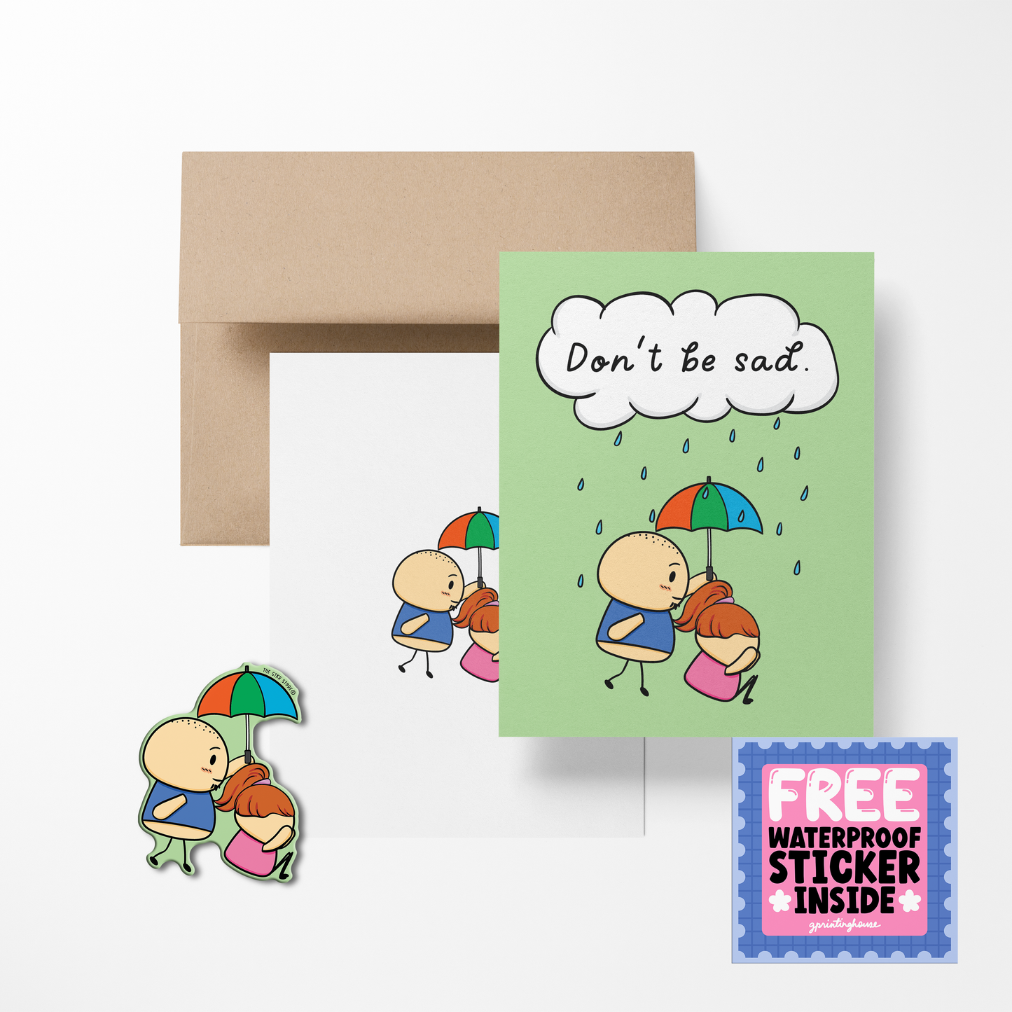 A greeting card with a cartoon character holding a umbrella, with the text 'Don't be sad' printed above. Accompanied by a brown envelope and a free waterproof vinyl sticker.
