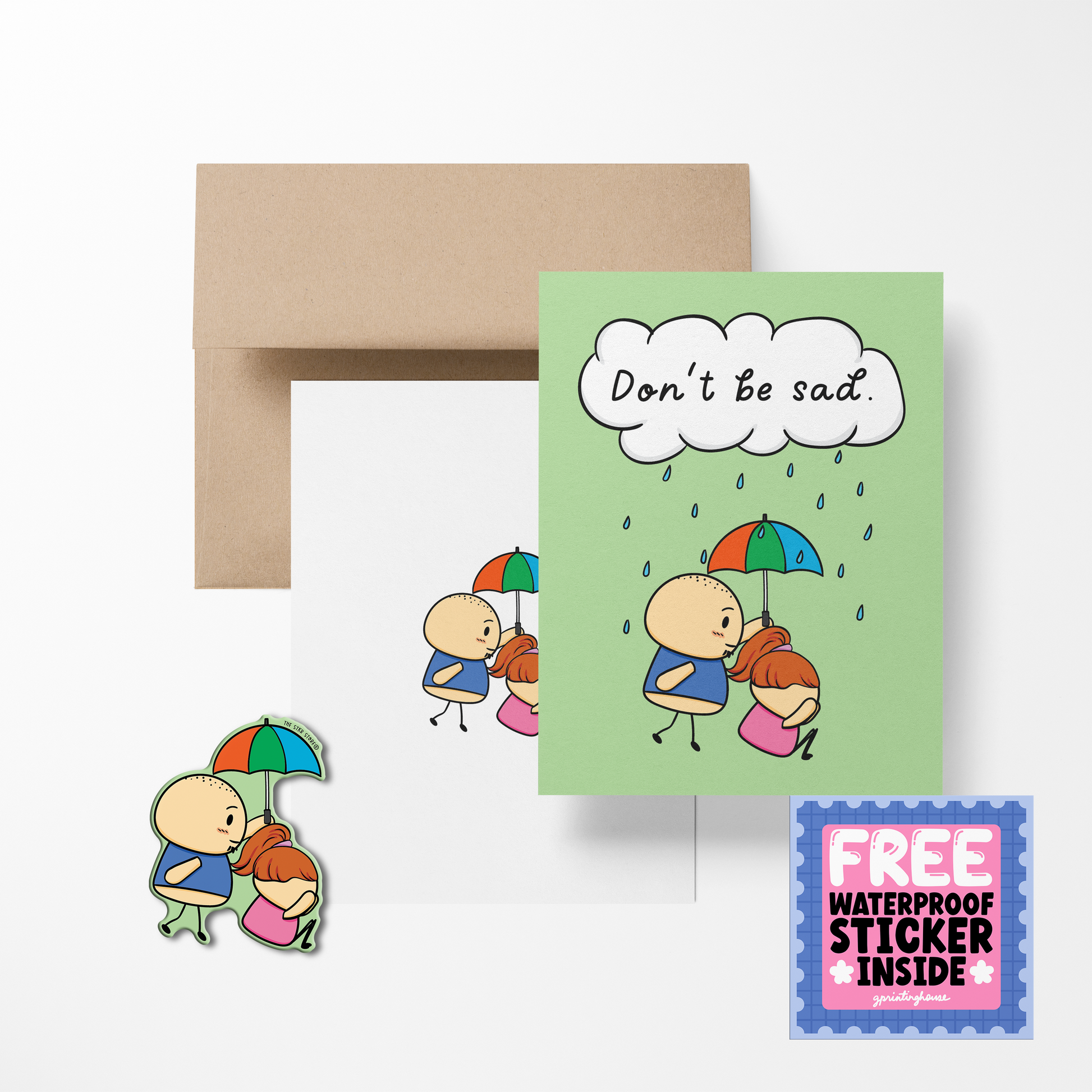 A greeting card with a cartoon character holding a umbrella, with the text 'Don't be sad' printed above. Accompanied by a brown envelope and a free waterproof vinyl sticker.
